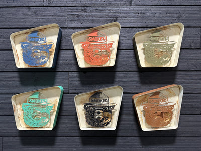 Handcrafted Smokey Bear metal signs in assorted vintage colors with rusted patina, mounted on a wooden wall – custom steel wall art for cabins or outdoor decor.